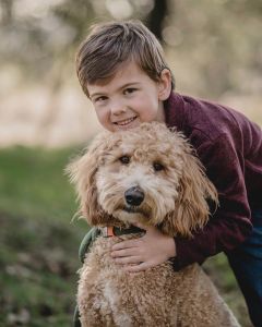 A young boy with brown hair, wearing a maroon jacket, hugs a fluffy, light brown Goldendoodle outdoors. Both are looking at the camera and smiling, showcasing why Goldendoodles Arizona are raised by some of the best breeders in the Southwest.